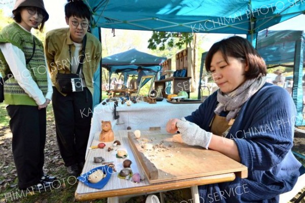 木彫りのアクセサリーを実演する後志管内ニセコ町の木工作家（右）。ナイフ１本で器用に形作る様子を来場者も興味深く見守る