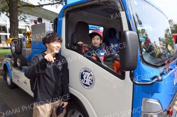 ＪＡＦの子ども用サイズの制服を着してレッカー車に試乗する子どもの姿に目を細める父親