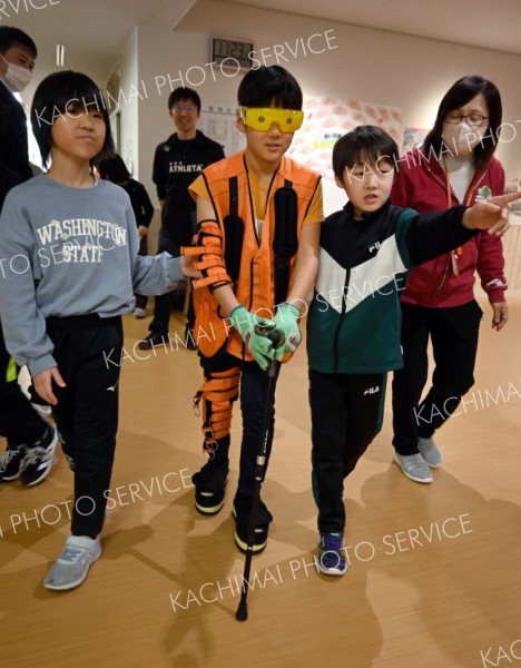 高齢者疑似体験セットを身に着け、クラスメートのサポートを受けながら歩く児童